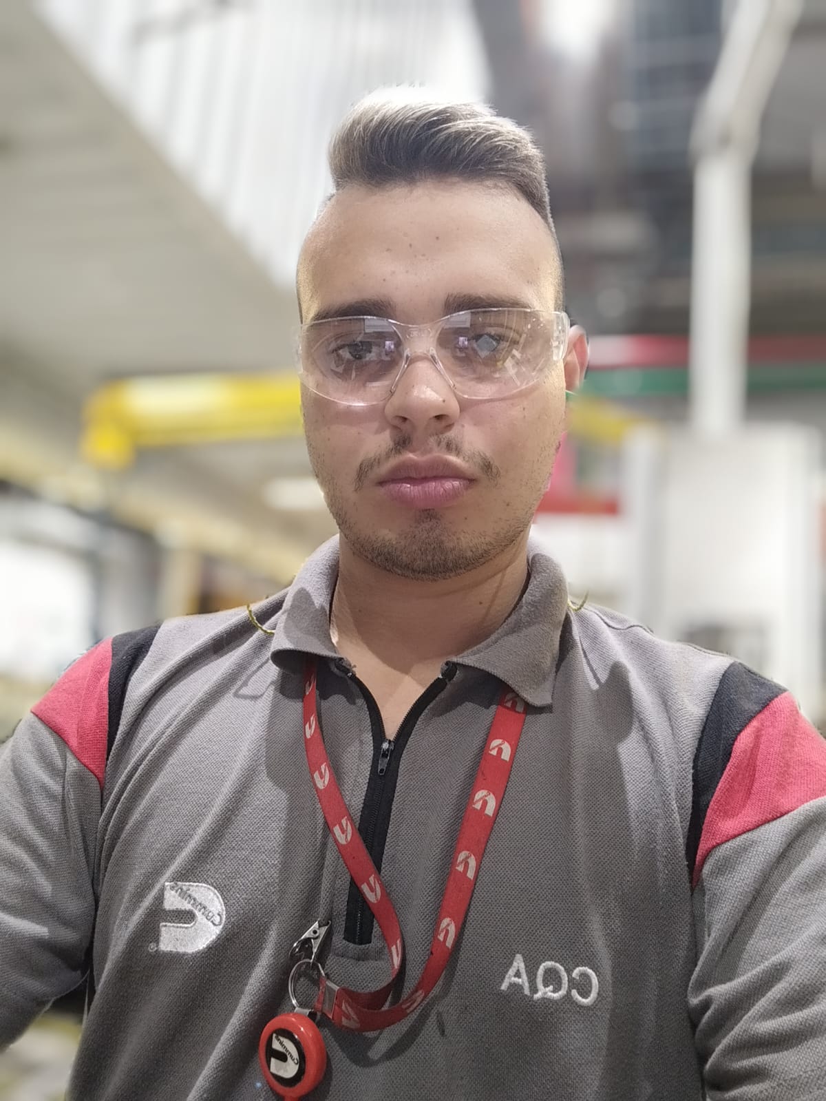 Um homem branco, de cabelos loiros e barba loira, utilizando óculos de proteção transparentes e camiseta cinza, posa para foto com feição séria. Ele tem crachá vermelho com detalhes brancos em volta do pescoço. 